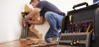 A picture focusing on a tool bag filled with a variety of tools while out of focus a worker performs repairs on plumping pipes behind a wall